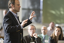 Photograph of a man standing and speaking to a crowd of people sitting